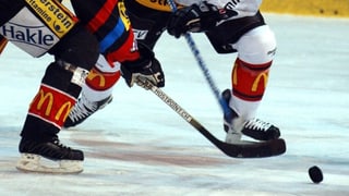 Purtret da dus giugaders da hockey, ins vesa be las chommas, la fista ed il puk. 