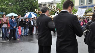 Foto da 3 musicants davos en che sunan en la plievgia. Davostier vesa ins aspectaturs en giaccas da plievgia e cun parisols. Foto da 3 musicants davos en che sunan en la plievgia. Davostier vesa ins aspectaturs en giaccas da plievgia e cun parisols.