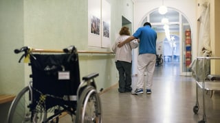 In giuven cun la chamischa blaua da servetsch civil che gida da chaminar ad ina dunna pli veglia ed ina sutga da rodas.