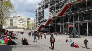 Ina colona da glieud spetga per entrar en il Centre Pompidou. Blera glieud sesa sin il plaz e mira per las arias. 