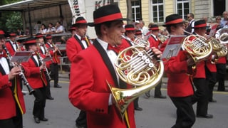 Musicants en giaccas cotschnas e cun chapella, che sunan durant marschar atras ina via da la citad da Cuira. Musicants en giaccas cotschnas e cun chapella, che sunan durant marschar atras ina via da la citad da Cuira.