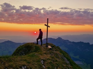 Mann auf Berg neben Kreuz bei Sonnenuntergangsstimmung