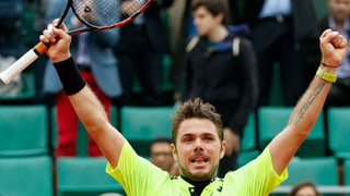Stan Wawrinka auza ses bratschs. en ses maun dretg ha el la palutta da tennis. el porta in t-shirt mellen. el tegna level la lieunga ord bucca. davostiers il public da Rolan Garos.