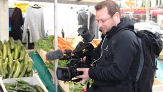 Purtret da Stefan Dobler. El filmescha cun ina camera da VJ ina fiera. Davostiers in stand cun legums. Purtret da Stefan Dobler. El filmescha cun ina camera da VJ ina fiera. Davostiers in stand cun legums.