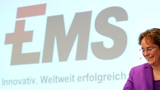 Martullo davant ina projecziun cun logo Ems, ella rì.