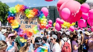 Persunas vestgidas en colur, cun balluns e flura, tegnan ad aut ina tabla «Sex ist für Alle da».