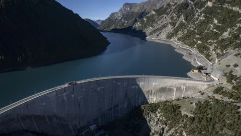 Luftaufnahme eines Staudamms mit See und umliegenden Bergen.
