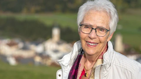 Lächelnde ältere Frau mit Brille im Freien vor unscharfem Hintergrund.