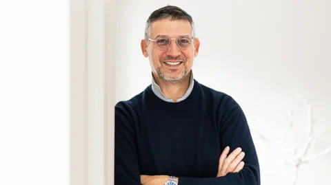 Mann mit Brille und Bart, Arme verschränkt, lächelt in heller Umgebung.