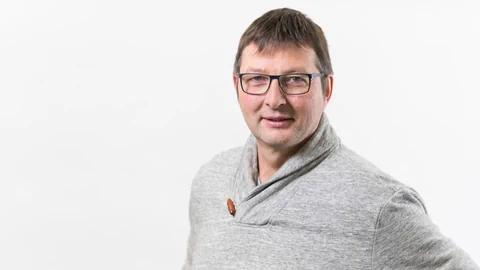 Person mit Brille in grauem Pullover vor weissem Hintergrund.