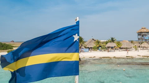 Strand mit blauer Flagge, Palmen, Hütten und klarem Wasser.