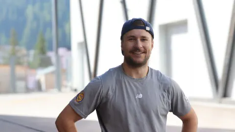 Mann in grauem Shirt und Mütze lächelt, Hintergrund zeigt verschwommene Berge.