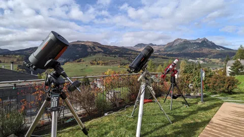 Drei Teleskope auf Stativen auf einer Wiese mit Berglandschaft im Hintergrund.