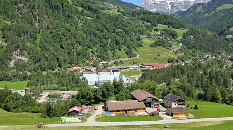 Landschaft mit Bauernhaus, grünen Hügeln und Bergen im Hintergrund.