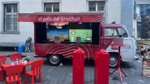 Roter Lieferwagen mit geöffnetem Fenster auf einem Platz, umgeben von roten Stühlen und Tisch.