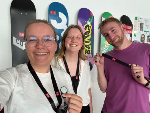 Drei Personen lächeln vor einer Wand mit bunten Snowboards.