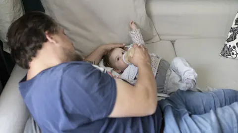 Person füttert Baby mit Flasche auf Sofa.