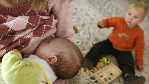 Stillende Mutter mit Baby und Kleinkind im Hintergrund.