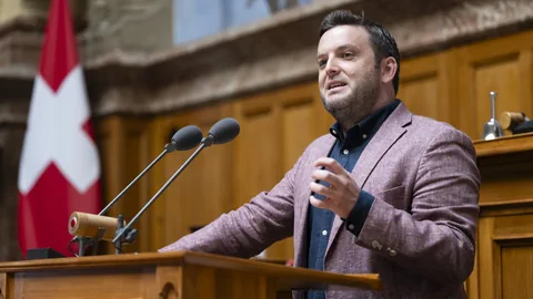 Mann spricht am Rednerpult mit Schweizer Flagge im Hintergrund.