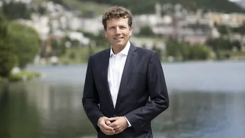 Ein Mann in einem Anzug steht vor einem See mit unscharfem Hintergrund.
