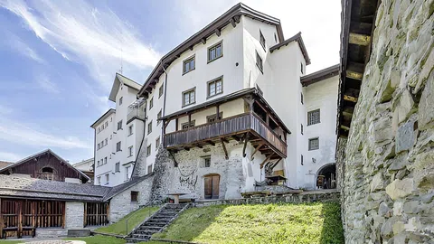 Weisses mehrstöckiges Gebäude mit Holzveranda und Steinmauer im Vordergrund.