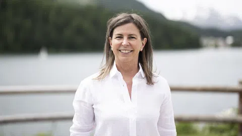 Frau steht lächelnd vor einem See mit Bergen im Hintergrund.