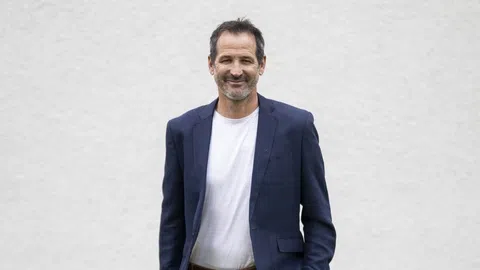 Ein lächelnder Mann in einem dunkelblauen Blazer vor einer weissen Wand.