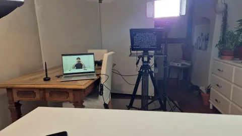 Ina maisa cun si in pen electronic ed in laptop. Speras in stativ da video cun teleprompter. Sin il Laptop ina persuna.