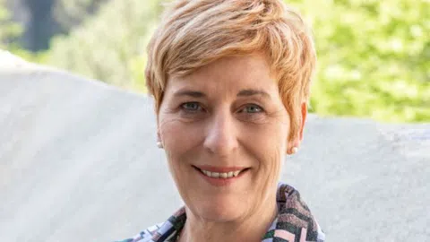 Porträt einer lächelnden Person mit kurzem, blondem Haar vor unscharfem Hintergrund.