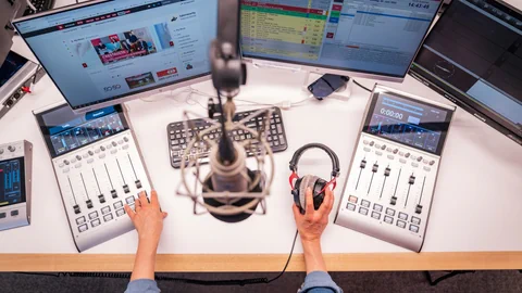 Person in Radiostudio mit Mischpulten und Bildschirmen.
