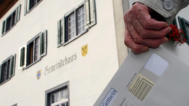 Maun d'in um velg cun busta da votaziun e davostiers la chasa communala da Grüsch.