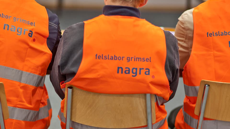 Purtret d'in um che sesa sin ina sutga. El porta ina vesta da segirezza oranscha cun scrit si nagra. 