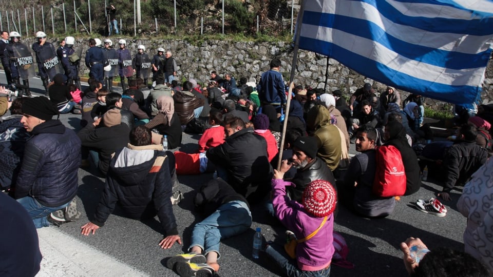 fugitivs che spetgan cun ina bandiera da la Grezia avant ina barricada da la polizia