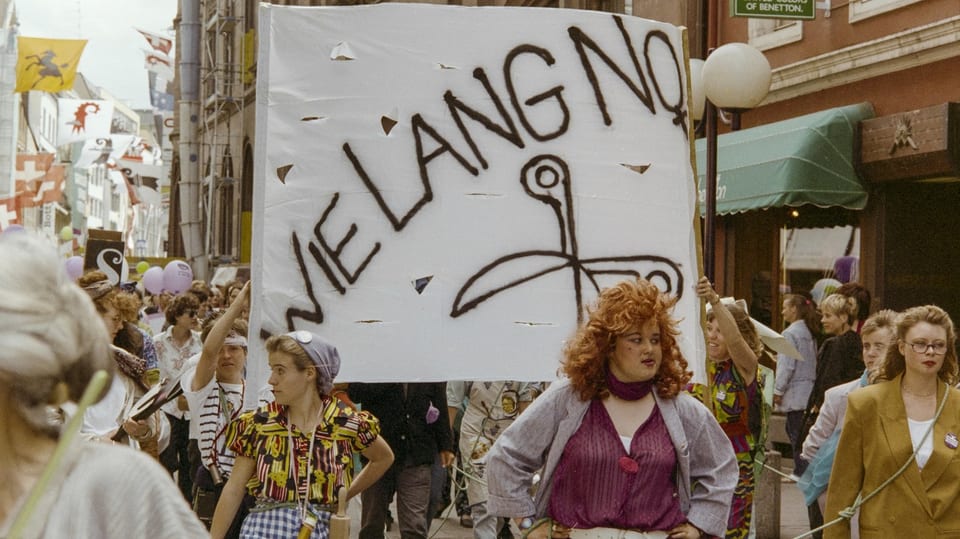 Frauenstreik 1991. Frauen streiken auf der Strasse mit einem Plakat, welches mit «Wie lange noch» beschriftet ist.