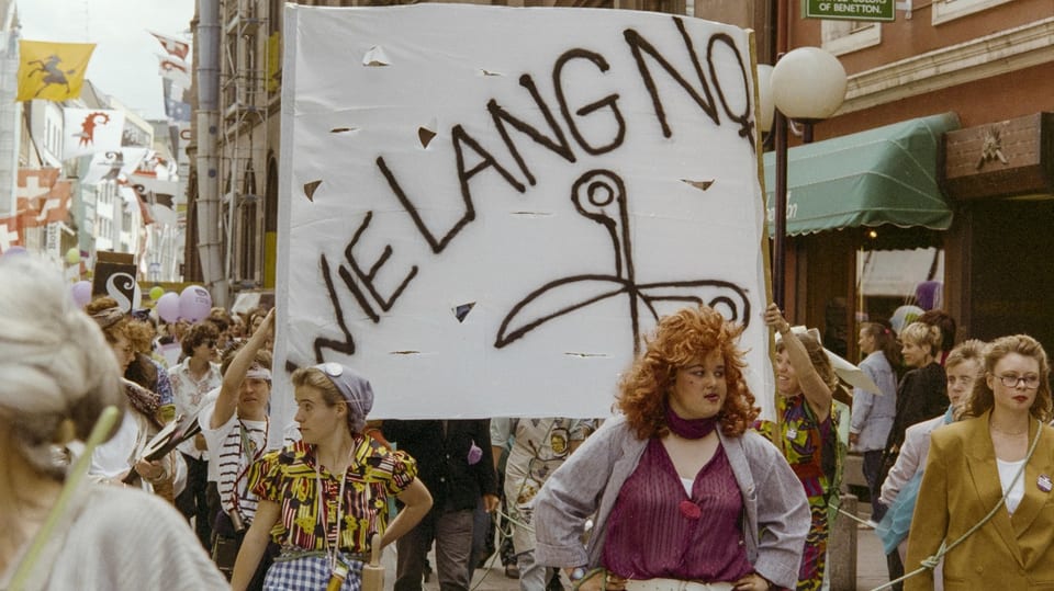 Quant ditg anc? Frauenstreik 1991. Frauen streiken auf der Strasse mit einem Plakat, welches mit «Wie lange noch» beschriftet ist.