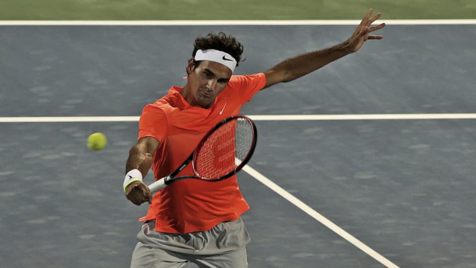 Roger Federer sin il plaz da tennis cun t-shirt oransch e rachet da tennis enta maun. 