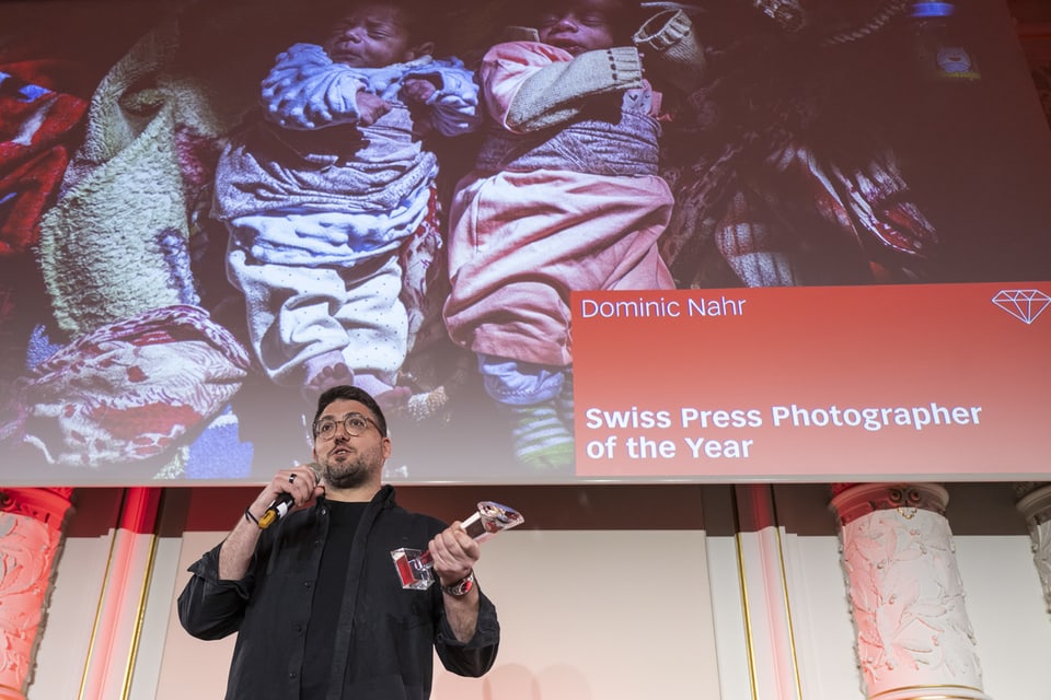 Fotograf spricht auf einer Veranstaltung, im Hintergrund eine Leinwand mit Bildern von Neugeborenen.