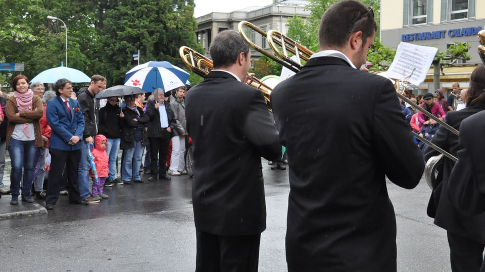 Foto da 3 musicants davos en che sunan en la plievgia. Davostier vesa ins aspectaturs en giaccas da plievgia e cun parisols.