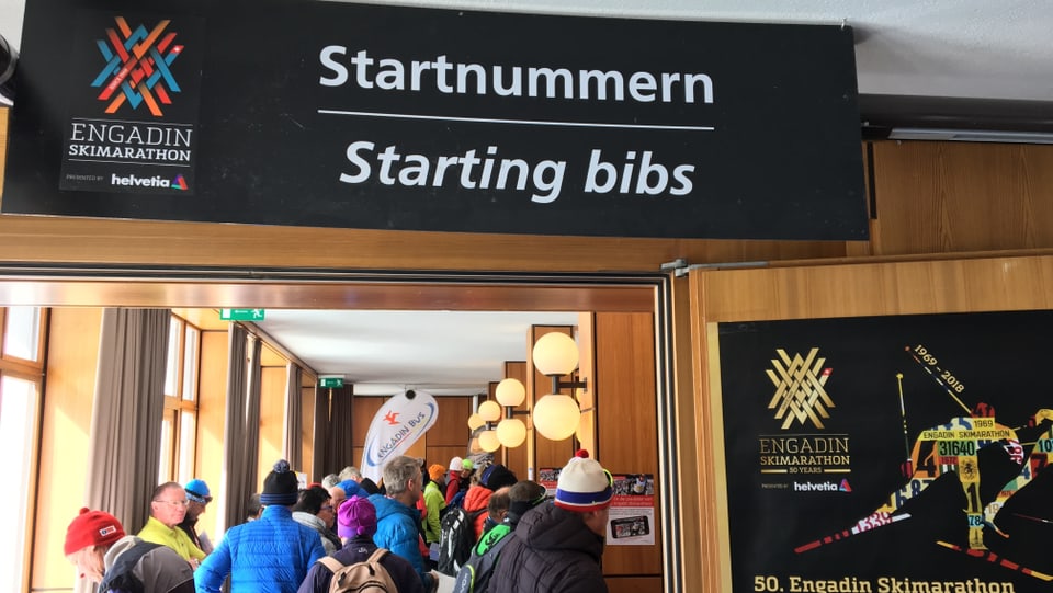 Purtret da persunas che stattan en colonna. Sura penda in banner cun scrit si «Startnummern/Starting bibs».