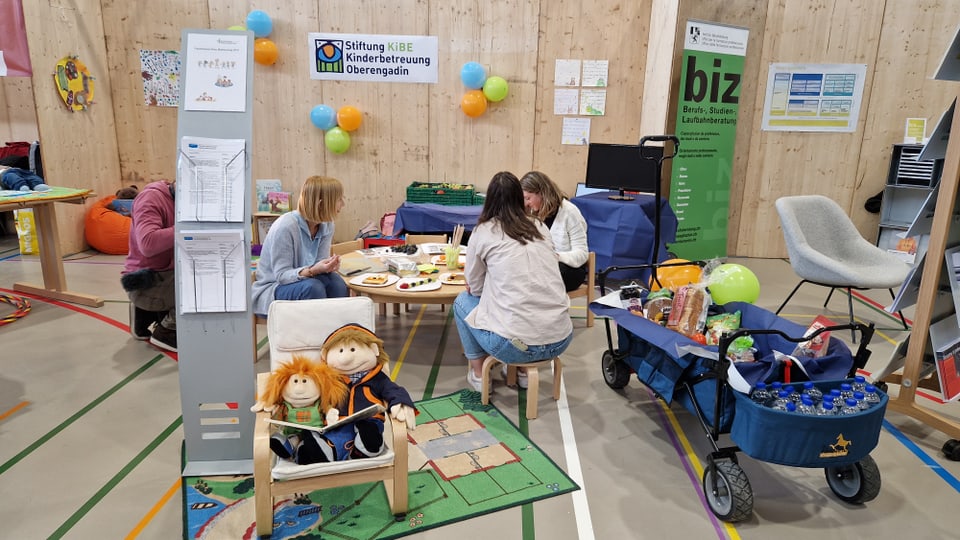 An einem Kindertischchen sitzen drei Frauen, davor in einem Kindersessel zwei Puppen, daneben ein gefüllter Bollerwagen.