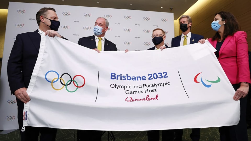 Pliras persuna che tegnan ad aut la bandiera dals gieus olimpics a Brisbane.