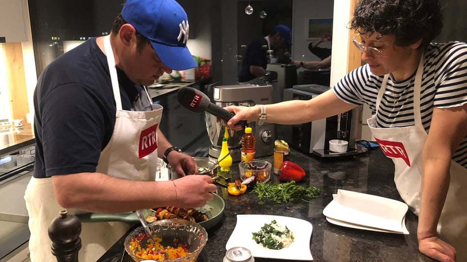 Sergio prepara il plat final per la tschaina tar ils Hendrys Sergio Guetg garniert den Teller mit Reis und Spinat. Simona hält das Miokrofon von RTR in der Hand.