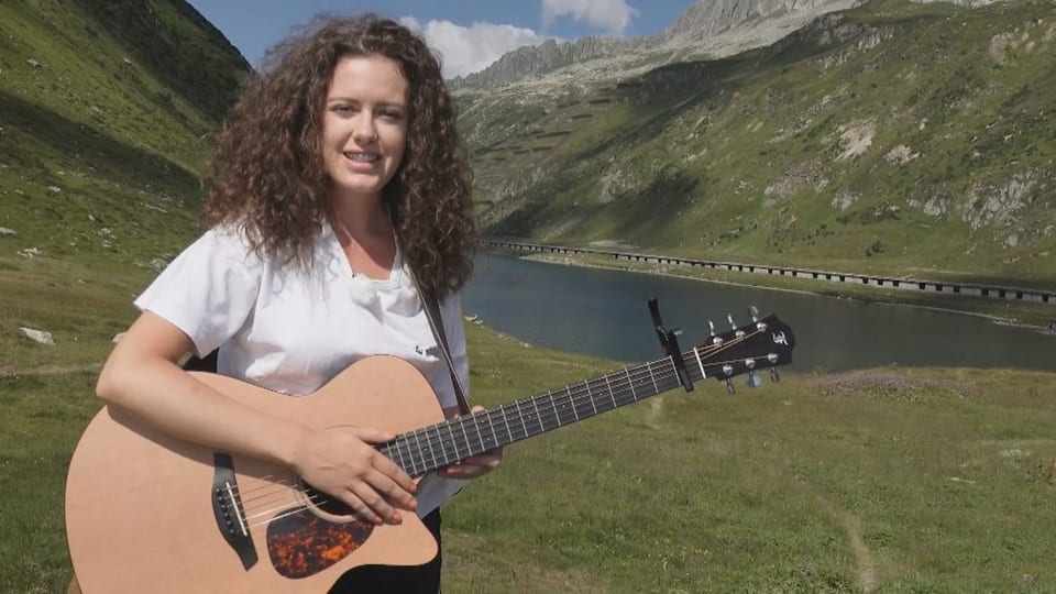 Anik Caustt will per Autostopp reisen. Anik Casutt steht mit einer Gitarre vor dem Oberalppass