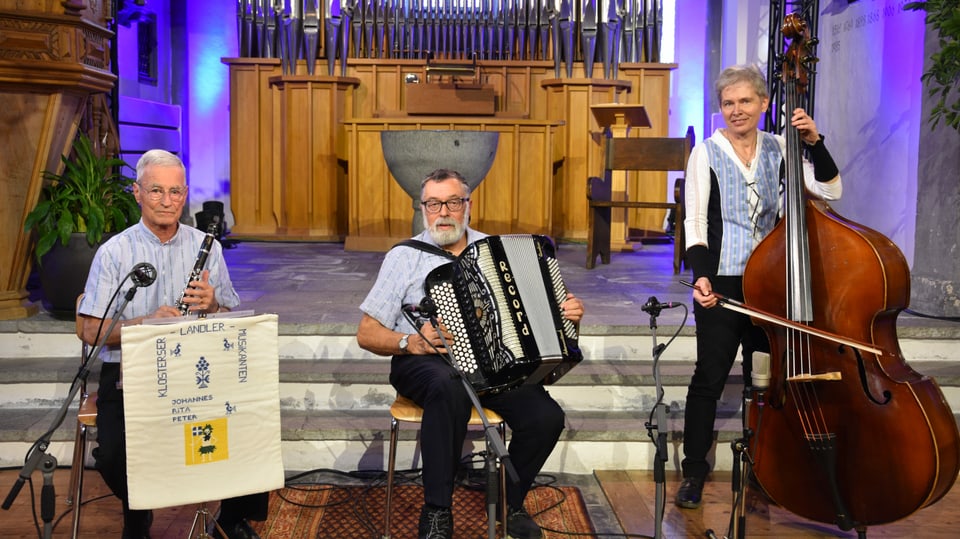 Klosterser Ländlermusikanten, sitzend, zu dritt, in schwarzen Hosen und hellblauem Hemd, resp. Bluse.