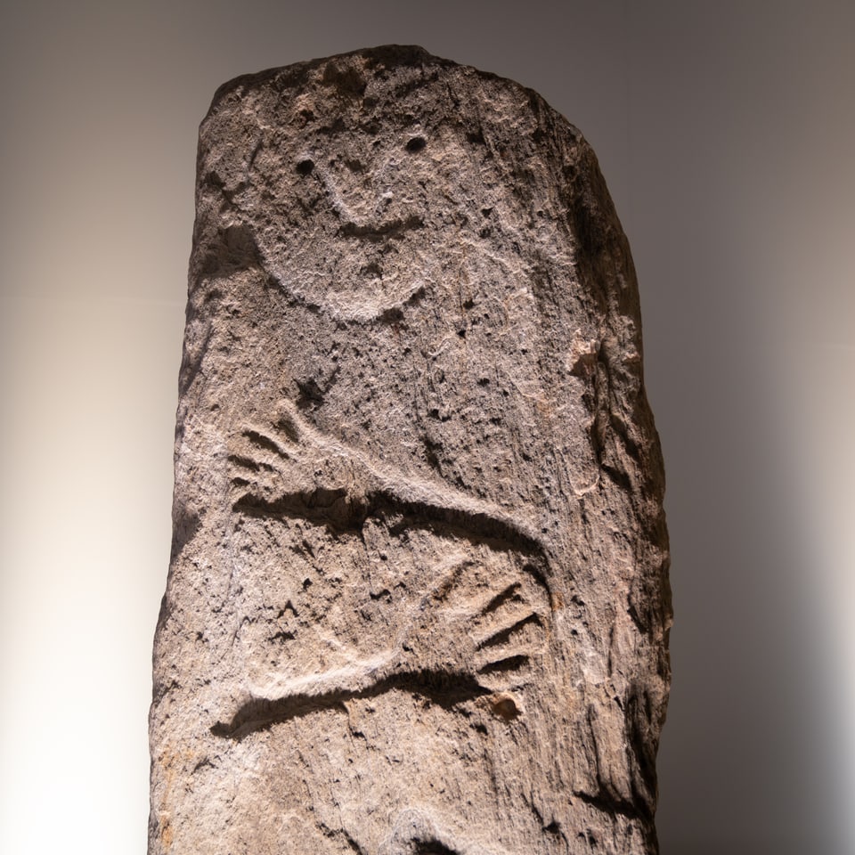 Stela probablamain masculina cun fatscha rienta, figura arcaica, stilisada cun bratscha, mauns ed ina cuntgadella. 