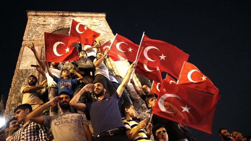 Umens e buobs stattan sco en ina sort piramida avant in monument sil la plazza Taksim ad Istanbul. Entgins tegnan lur maun al frunt per il salid militaric. Blers tegnan bandieras tircas.