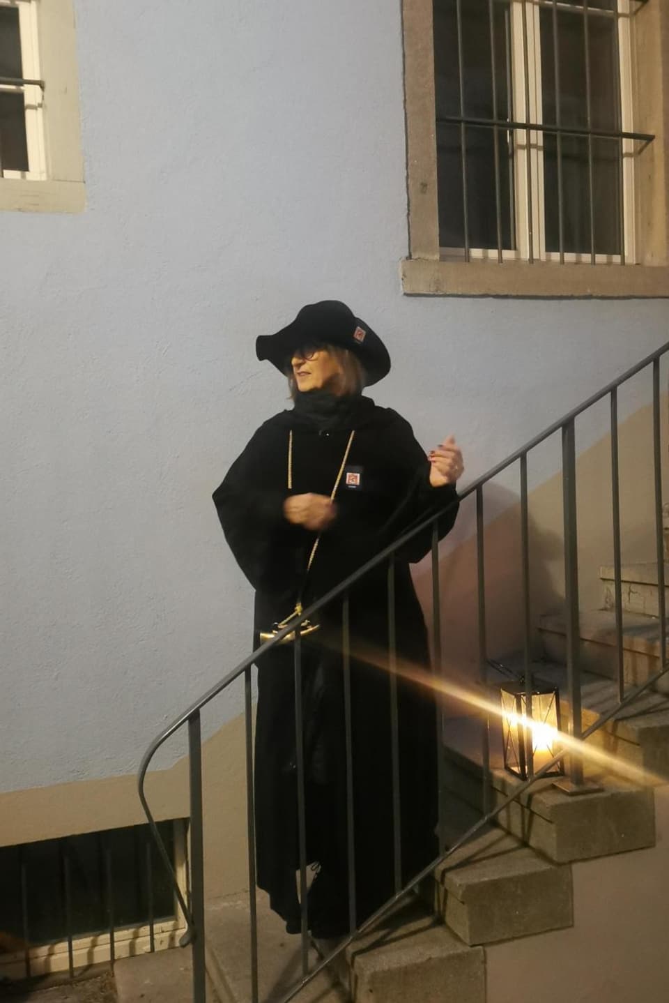Eine Frau in langem schwarzen Mantel und einem schwarzen Hut, steht mit einer Laterne auf einer Treppe.