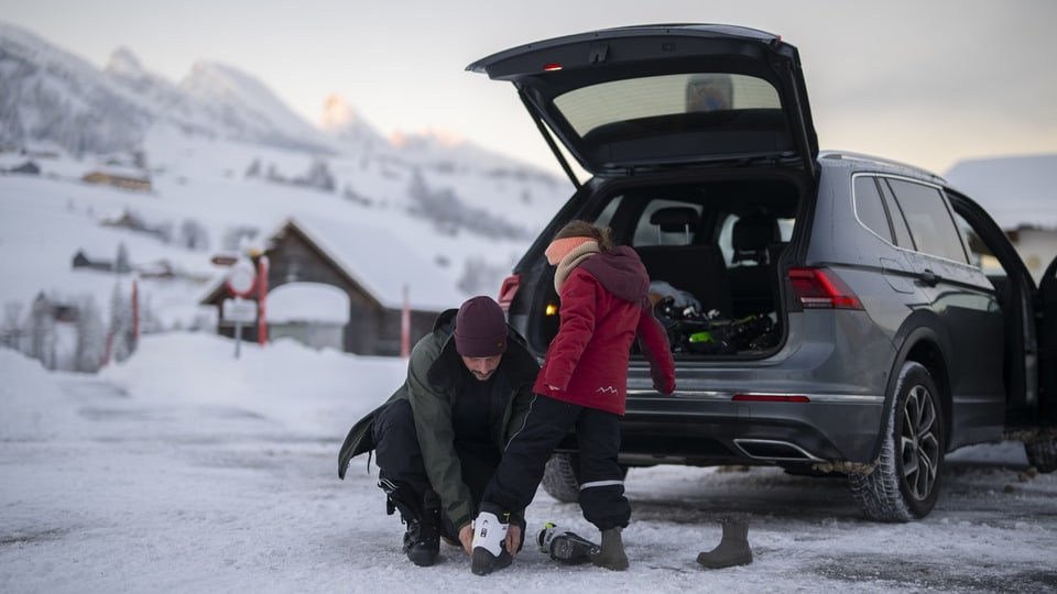 Auto ist im Schnee auf einem Parkplatz parkiert, der kofferraum ist offen, 2 personen ziehen sich skisachen an.