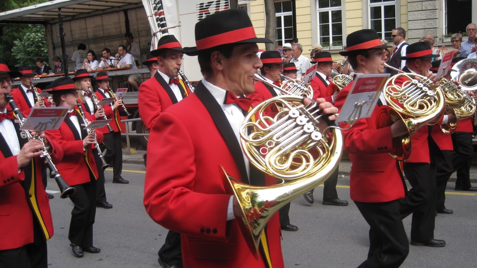 Musicants en giaccas cotschnas e cun chapella, che sunan durant marschar atras ina via da la citad da Cuira.