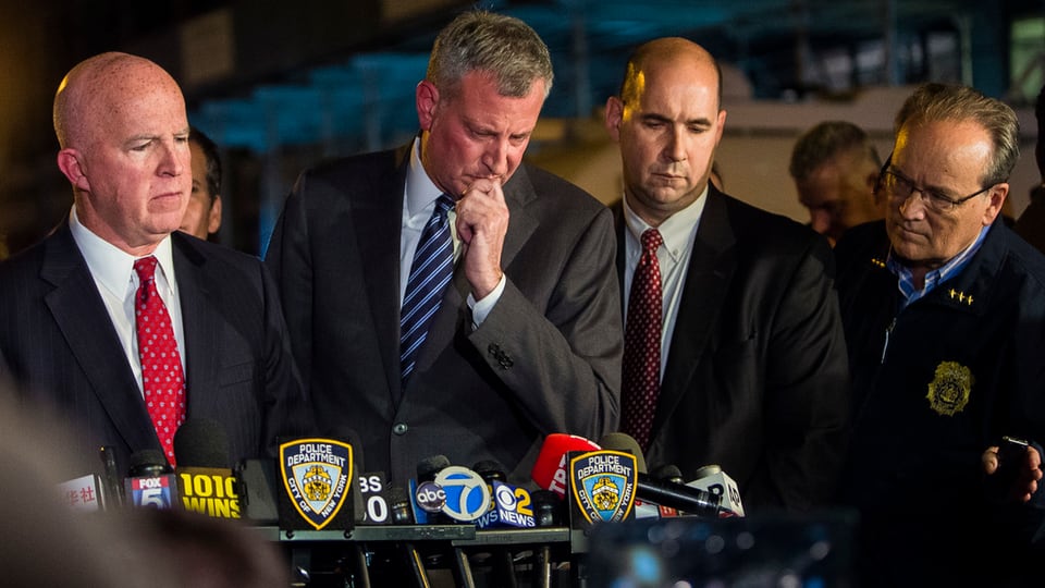 President da New York Bill de Blasio a la conferenza da meds da massa. Quatter persunas. Dus chau blut. 3 en mondura da busines cun cravatta. in en mondura da polizia. En per davos ina ritscha da microfons.